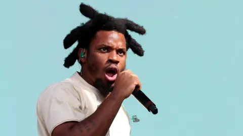 Getty Images Denzel Curry has thick black hair styled on each side of his head. He wears a white t-shirt, a green ear-piece and holds a black microphone as he performs against a turquoise background. He has a tattoo on his forearm.