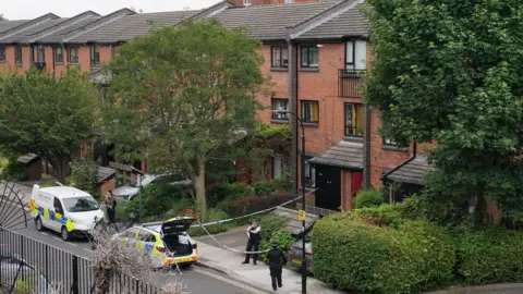PA A blue and white police cordon set up outside the flat in London, with several police officers in uniform monitoring the perimeter. There are two police vehicles parked beside the pavement. The buildings are made from red brick with black Juliette balconies and grey slate roofs.