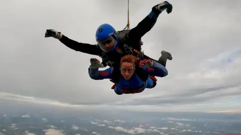 Kim Cook Two people strapped together in the sky, skydiving. They both have red, blue and black flight suits on with goggles and have there arms out.