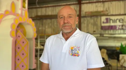 Martyn Maguire pictured in a Battle of Flowers shed. A float is partially visible in the background. He has buzzed hair and a stubble beard. He is wearing a white polo shirt.