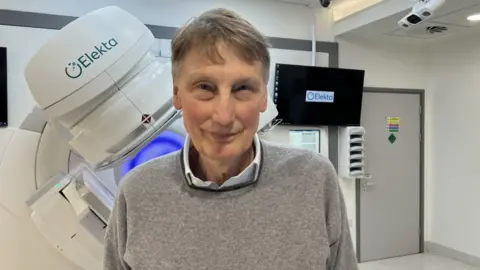 Prof Adrian Crellin wearing a grey jumper and blue shirt stood in front of cancer treatment equipment at a hospital 