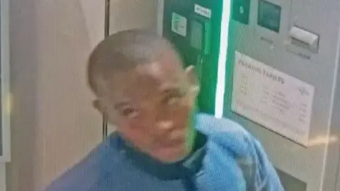 A dark skinned man with a shaved head and wearing a blue top is seen on CCTV, turning away from a parking ticket pay point