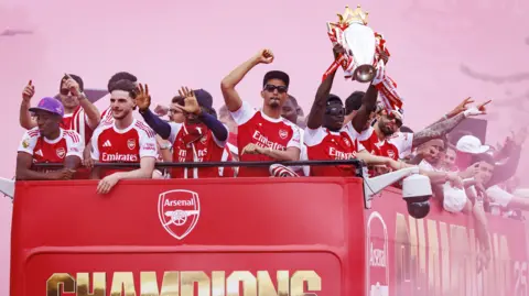 Bukayo Saka of Arsenal (R) lifts the English Premier League trophy as he and teammates ride an open-top bus during Arsenal Champions Parade in north London