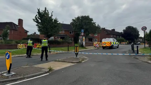 BBC/ Emily Johnson Two police officers, with their backs to the camera, guard a police cordon blocking off a residential street. A police van is within the cordon. 