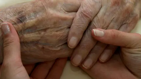The image shows a close-up of two hands gently holding an older person’s hand. The older hand appears wrinkled and has visible veins and age spots, suggesting advanced age. The other hands look younger and are positioned in a supportive, caring way, cradling the older hand from underneath and around the sides.