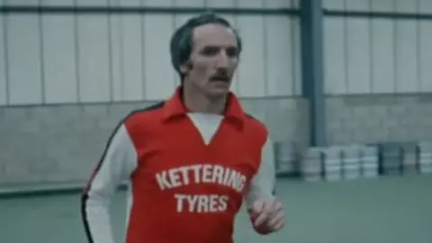 Derek Dougan, who has a moustache and receding hairline, is wearing a red football shirt with white sleeves. "Kettering Tyres" is emblazoned across the front in white capital letters. He appears to be training in a large sports hall and there are what appear to be beer kegs on the wall behind him.