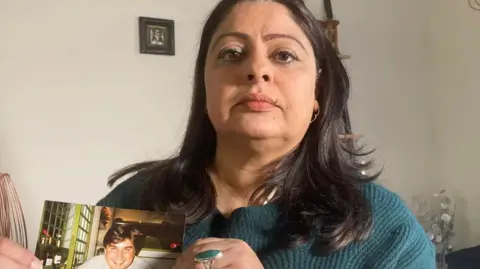 Kam Miller Kam is holding a photograph of Neil. She is pictured at home and has long brown hair and is wearing a green cardigan. 