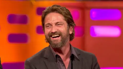 Gerard Butler laughs whilst looking to the left, we can only see from the shoulders up. He has a longer hair down to just under his ears. He has a beard and wears a black suit jacket with a purple shirt and the top buttons are open. 
