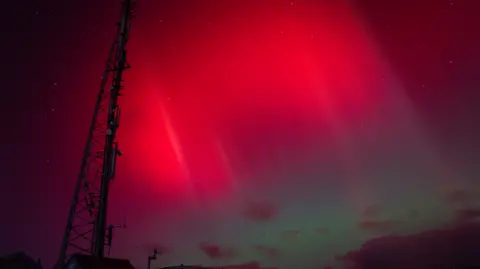 Chris Kirkham A telecoms tower stands in the foreground, backlit by bright red shafts in the night sky. Dappled clouds float in deep blue and green sky to the right,