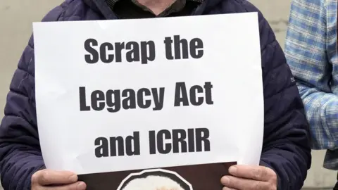 PA Media A person wearing a navy, padded coat holds up a sheet of white paper with 'Scrap the Legacy Act and ICRIR' in black font.