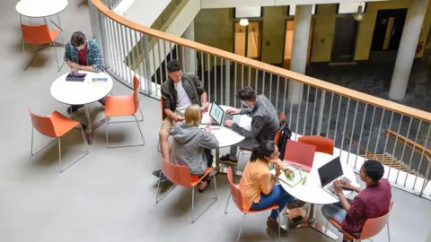 Getty Images Students sit on three tables. they sit at their laptops doing work. the pictures it taken from a high angle.