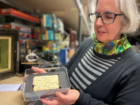 Leeds Musuems A woman is holding a plastic display box containing a broken, beige-coloured biscuit with faded blue handwriting on the surface of the biscuit. The woman is wearing a black cardigan, a striped top and a colourful scarf around her neck. In the background are shelves stacked with various items.