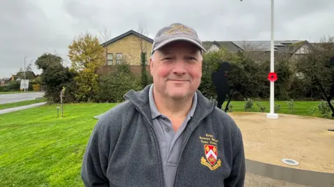 Tony Jones looks into the camera while standing in front of the memorial. He wears a grey cap and fleece, which both have the town council logo.