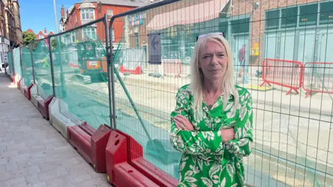 Tracy Jones is stood in the street next to large metal fencing with roadworks behind. She has long hair and is wearing a green and white floral shirt and has her arms folded.
