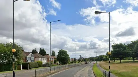 A Google Streetview screenshot of Normanby Road in Ormesby, where the crash happened. The road has two lanes going in one direction and a single-lane in the other. There is a green space on one side of the road and houses on the other and cars on the road in the distance.