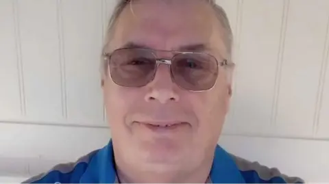 BBC David Blakey stands against a white wall wearing a blue and grey striped top and aviator-style glasses