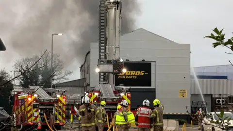 Paul Wood A ground level shot of fire crews wearing hard hats and holding hoses, next to the building which is on fire.