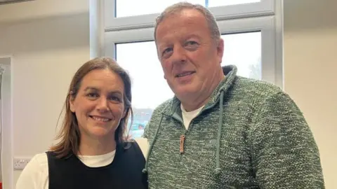 Richard and Andrea stand shoulder to shoulder. Richard has short grey hair, and wears a light green jumper and Andrea has shoulder length brown hair and wears a white t-shirt with a black knitted vest over the top.