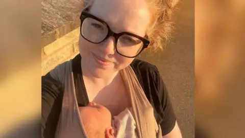 Laura Ayoade A woman with blonde hair and black glasses smiles at the camera. She is carrying a baby in a sling on her front.