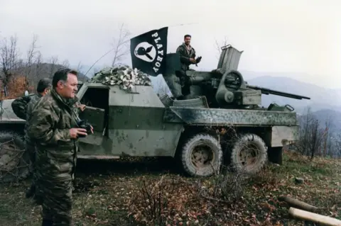 Gamma-Rapho Images via Getty Images General Mladić leads his troops in 1994. He is standing next to a military vehicle and he is holding binoculars 