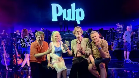 The band Pulp are sat on a stage holding their award from the Official Charts