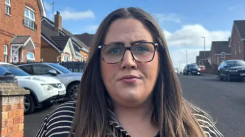 A woman looking at the camera, she has dark hair, glasses and is wearing and black and white shirt