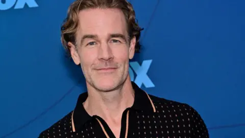 Getty Images James Van Der Beek wears a black polo and stands in front of a blue background