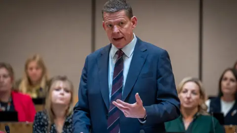Senedd Cymru Rhun ap Iorwerth speaking while gesticulating in a blue suit, and light blue shirt, and tie