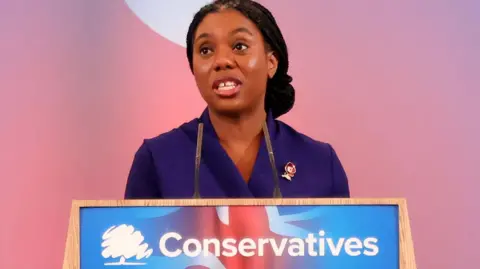 EPA Kemi Badenoch stands in front of a plinth bearing the Conservative logo. She is wearing a purple dress.