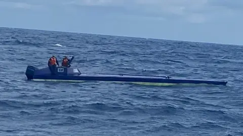 Two men wearing orange life vests and caps stand on top of a semi-submersible vessel seized by the Colombian Navy.