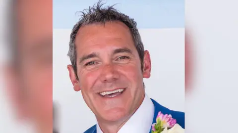 Jeremy Clark smiles at the camera. he is wearing a suit and white shirt with a floral buttonhole on his jacket.