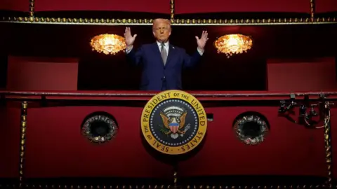 Hulton Archive via Getty Images Donald Trump with hands raised stands behind the presidential seal in the red presidential box at the centre