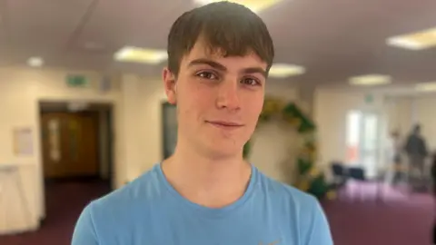 Fynn Stigant wears a blue t-shirt in a classroom. He has short dark hair and smiles.