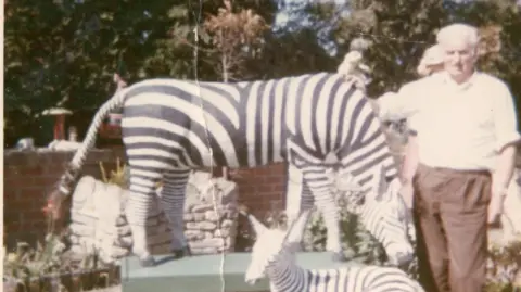 James and Christina Beveridge An old picture with a man in brown trousers and a white shirt with grey hair next to two statues of zebras