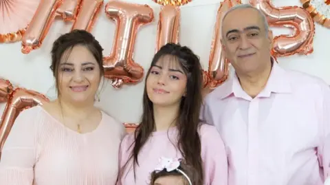 Pierre Naoum Carla (middle) standing with her mother (left) and father (right), dressed all in pink with rose gold lettered balloons behind them