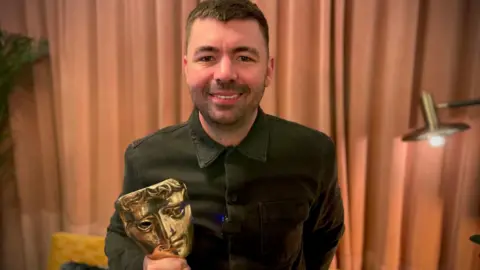 BBC Rich Pepiatt has short dark hair and stubble and is wearing a dark long-sleeved shirt buttoned to the top. He is smiling and holding the Bafta he won. Behind him are salmon coloured curtains