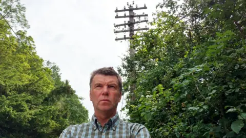 Martin Evans Martin Evans, with short brown hair and wearing a blue and brown checked shirt is standing in front of a telegraph pole, staring ahead in the distance. There are trees either side of him