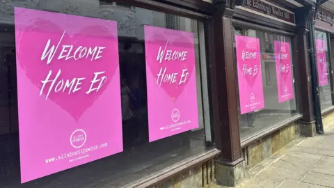 An empty store front with pink posters in the windows that read Welcome Home Ed.