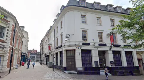 A view of a white multi-storey building on the corner of a street, next to a pedestrianised walkway.