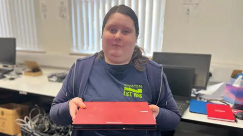 A young woman looks into the camera as she holds a red laptop computer. She has dark hair, tied back, and wears a blue T-shirt, with a green Giroscope logo, and a blue hoodie.