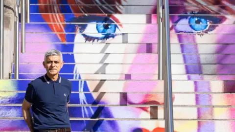 Mayor of London Sadiq Khan stands on steps painted with Taylor Swift's face. Her blue eyes loom above the mayor, who is striking a casual pose, dressed in a dark blue Fred Perry polo shirt. 