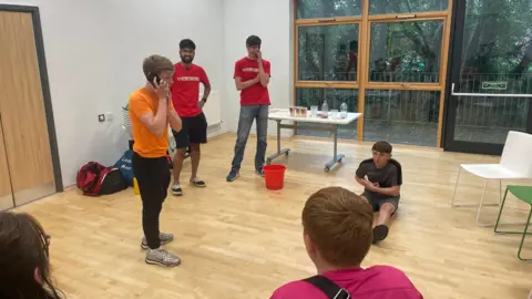 Two men in red StreetDoctors' T-shirts standing in a room looking at a boy in an orange T-shirt on a phone and a boy in a grey T-shirt and shorts on the floor holding his side