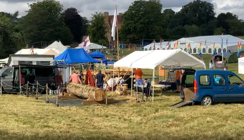 Luke Deal/BBC FolkEast visitors carry items from parked cars into the festival tents. A row of colourful flags can be seen behind the tents. 