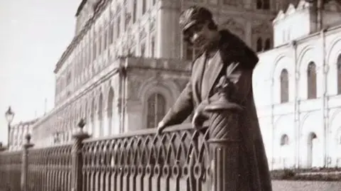 A black and white photo of Claire Sheridan in the Kremlin in Russia. The photo is quite blurry due to its age. Ms Sheridan wears a hat and a long trench coat. She is holding onto a gate outside of the Kremlin building.