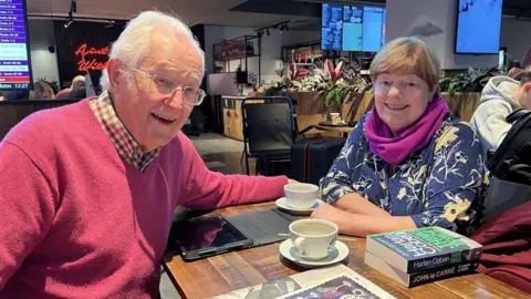 Iver and Margot Morton sitting at a table with cups of coffee, books and a tablet on the table. Iver has grey hair and glasses and is wearing a dark pink jumper. Margot has short brown hair and is wearing a blue top with a floral pattern and a purple scarf.