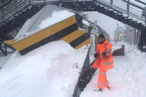 Network Rail Scotland A train snowplough covered in snow, with a worker in bright orange overalls next to it.