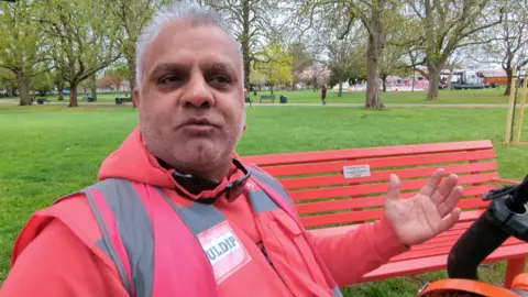 A man in a pink high visibility jacket stand in front of Bill's red bench in the park.  