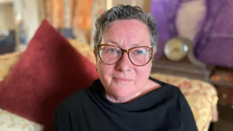 Emma Glasbey/BBC Dr Jacqui Lovell - a woman with short grey hair wearing glasses and a black jumper, sitting on a sofa.