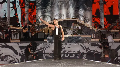 Getty Images Robbie Williams standing on stage at Knebworth holding a mic stand in one hand
