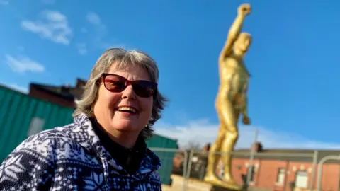 The woman in the hair smiling and posting in front of the huge golden statue of Geréntaya already has short hair on her head and the dust-effect is raised in the air. It was born with a male but it was removed to the background in this shot. The blue sky and rooftops can be seen behind the big statue. 